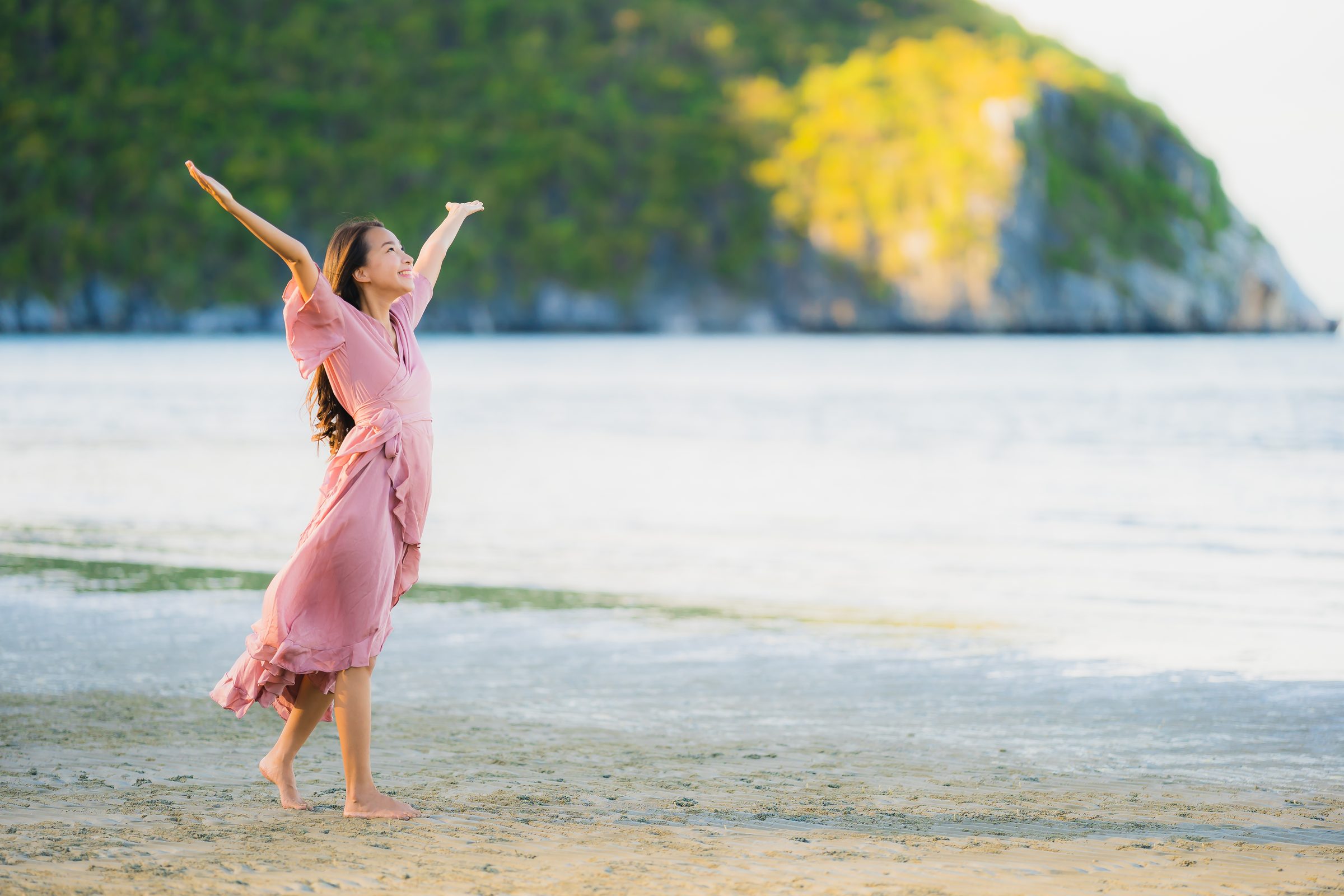 A woman in a pink dress on a beach in Thailand, her arms raised to the sky